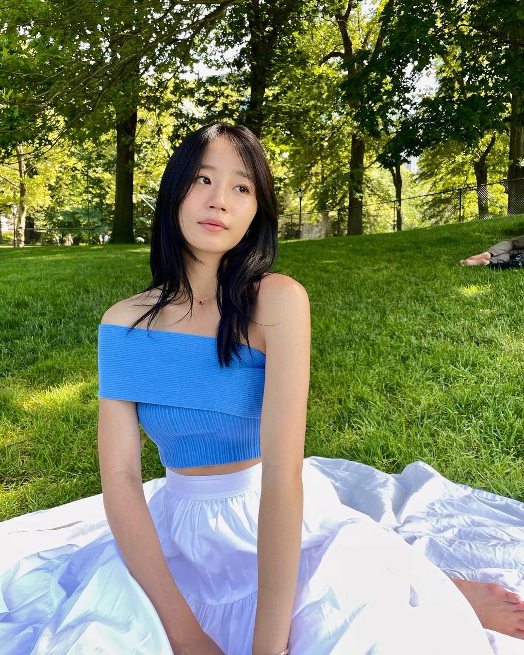 Gen Z - cô gái châu Á tóc đen dài xõa thẳng, mặc áo hở vai màu xanh dương và ngồi trên thảm cỏ